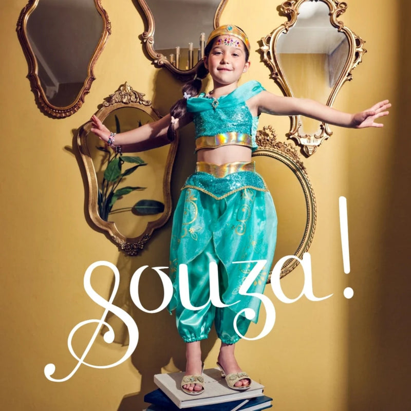 A young girl stands with arms outstretched wearing a turquoise and gold genie-style costume, posed in front of a yellow wall with ornate mirrors. Large white script text reading 'souza!' overlays the lower portion of the image.