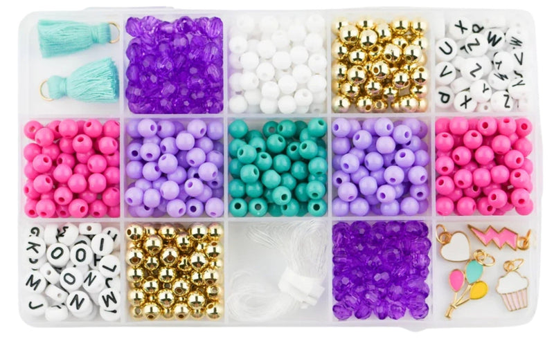 A clear plastic organizer tray filled with assorted craft beads and small charms on a white background. The compartments contain pink, purple, teal, white, and gold beads, along with letter charms, tassel ties, and small decorative charms.