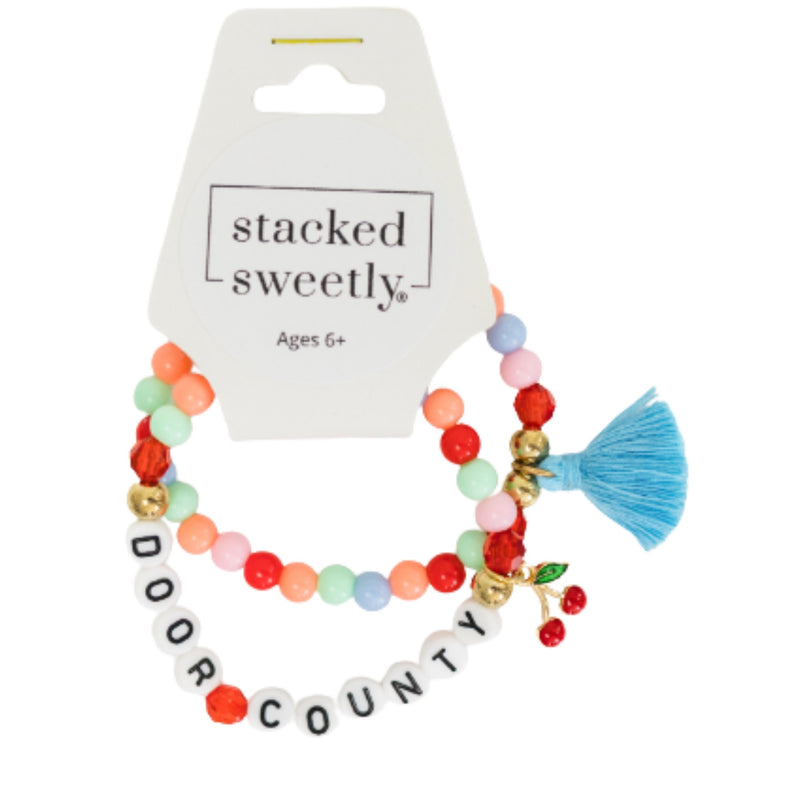 A two-piece beaded stretch bracelet set from stacked sweetly, featuring a multicolor bead bracelet and a white letter bead bracelet spelling 'DOOR COUNTY' with a blue tassel charm, displayed on a white retail card.