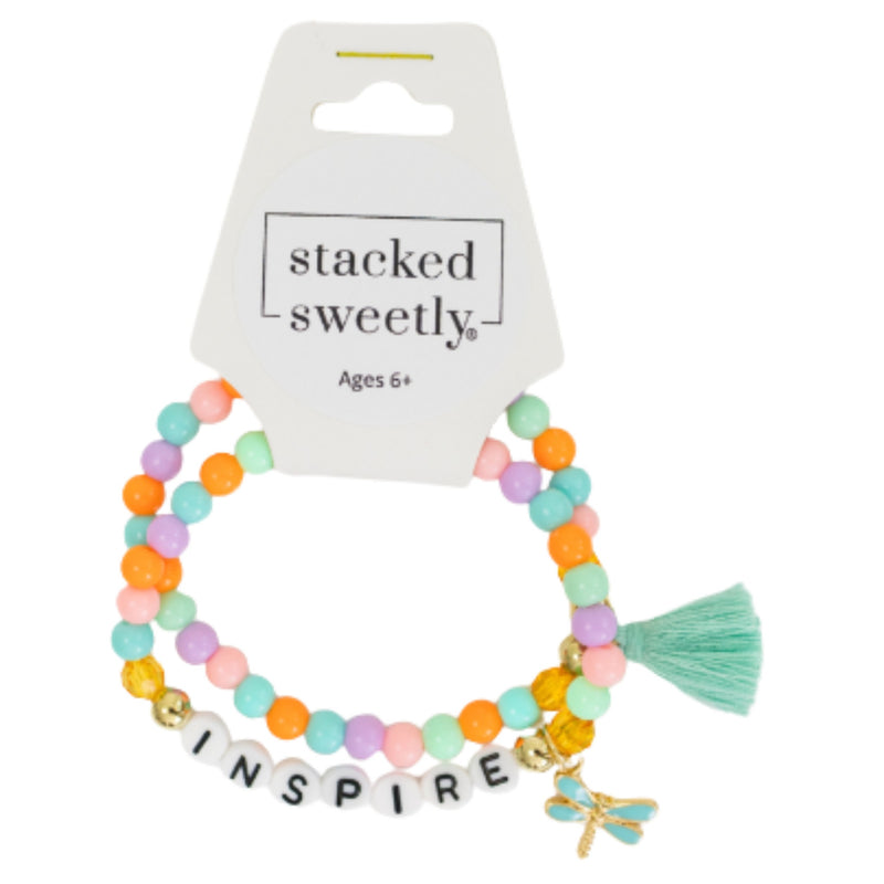 A two-pack of pastel beaded stretch bracelets by stacked sweetly, displayed on their white cardboard hanger. The bracelets feature pastel orange, mint green, lavender, and pink beads, a mint green tassel, white letter beads spelling 'INSPIRE', and a small gold dragonfly charm.
