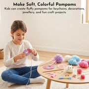 A young girl sits cross-legged on a cream rug, smiling while making a purple pom-pom with yarn. A small round wooden table to her right holds colorful craft supplies including pink, blue, purple, white, and yellow yarn, pom-poms, scissors, and a small plush toy.