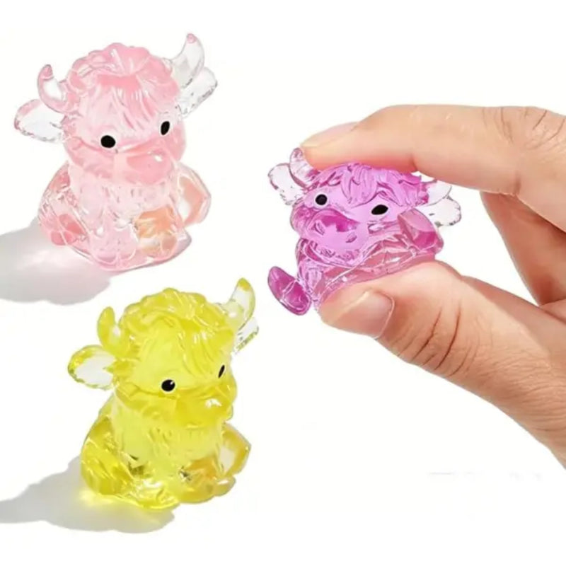 Three small translucent Highland Cow squishies in pink, yellow, and purple, with a human hand holding one purple cow on a white background.