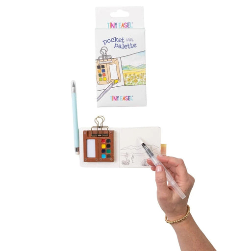 A hand holding a pen next to a miniature pocket paint palette with a watercolor wheel, a small paintbrush, and a drawing pad. The product packaging labeled 'TINYFASEER pocket palette' is shown above on a white background.