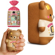 Kumapan Loafie Bear squishy toy shown in its pink packaging, held in a hand to demonstrate its soft squeezable texture, and standing upright on its own. The bear is light brown with a cream shirt and yellow patch on its forehead.