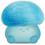 A light blue silicone facial cleansing brush with a rounded, squishy base and a dense, textured bristle head, featuring a simple smiling face on the front.