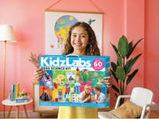 A young girl with curly brown hair stands in a pink room holding a large blue box for the KidzLabs Mega Science Kit. The box features colorful illustrations of science experiments and tools, and the brand name 'KidzLabs' is clearly visible.