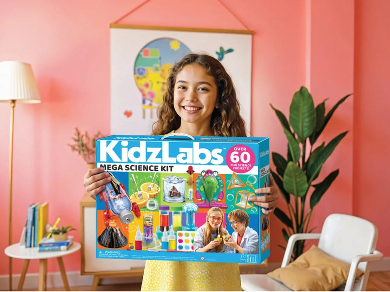 A young girl with curly brown hair stands in a pink room holding a large blue box for the KidzLabs Mega Science Kit. The box features colorful illustrations of science experiments and tools, and the brand name 'KidzLabs' is clearly visible.