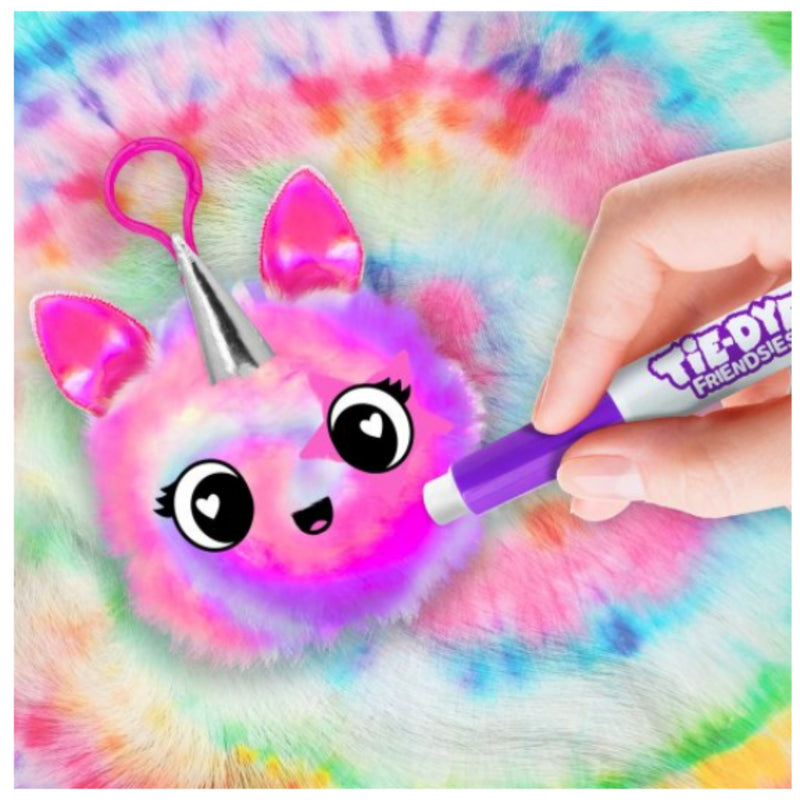 A close-up of a fluffy pastel tie-dye keychain or bag charm being dyed with a purple marker. The fuzzy item has pink ears, large cartoon eyes, and a silver horn, set against a matching tie-dye background.