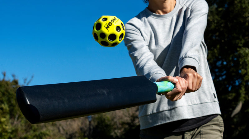 Person holding black cricket bat with yellow moon ball in outdoor setting