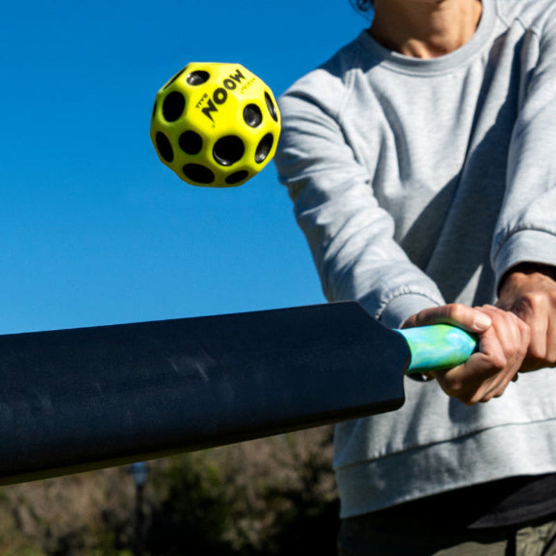 Person swinging black foam bat at yellow Waboba Moon Ball outdoors against blue sky