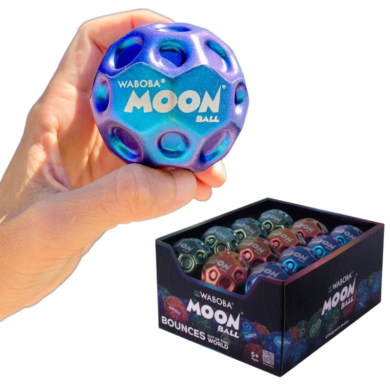 A hand holds a blue and purple iridescent Waboba Moon Ball, with a black display box of multiple colored balls below on a white background.