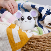 Cute white mouse-shaped toy with purple ears hiding in a basket of colorful cloths.