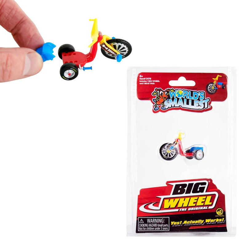 A hand holds a miniature red, yellow, and blue tricycle (Big Wheel) toy above its retail packaging on a white background. The package reads 'World's Smallest Big Wheel' and shows the toy inside a clear blister pack.