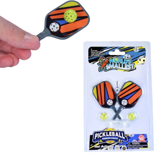 A hand holds a small pickleball paddle above its retail packaging on a white background. The paddle has orange, black, and yellow striped wings with a gray handle, and the package shows World's Smallest Pickleball with two matching paddles and yellow pickleballs.