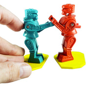 Two small plastic toy robots standing on yellow bases, with one robot in teal being held by a human hand and the other in red standing on a yellow base against a white background.