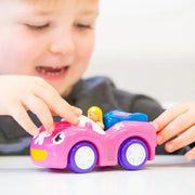 A young child's hands and face are visible as they play with a pink toy convertible car with purple wheels on a white surface.