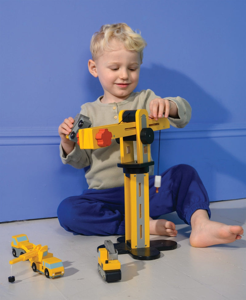 Child playing with yellow wooden crane toy and construction vehicles on blue background