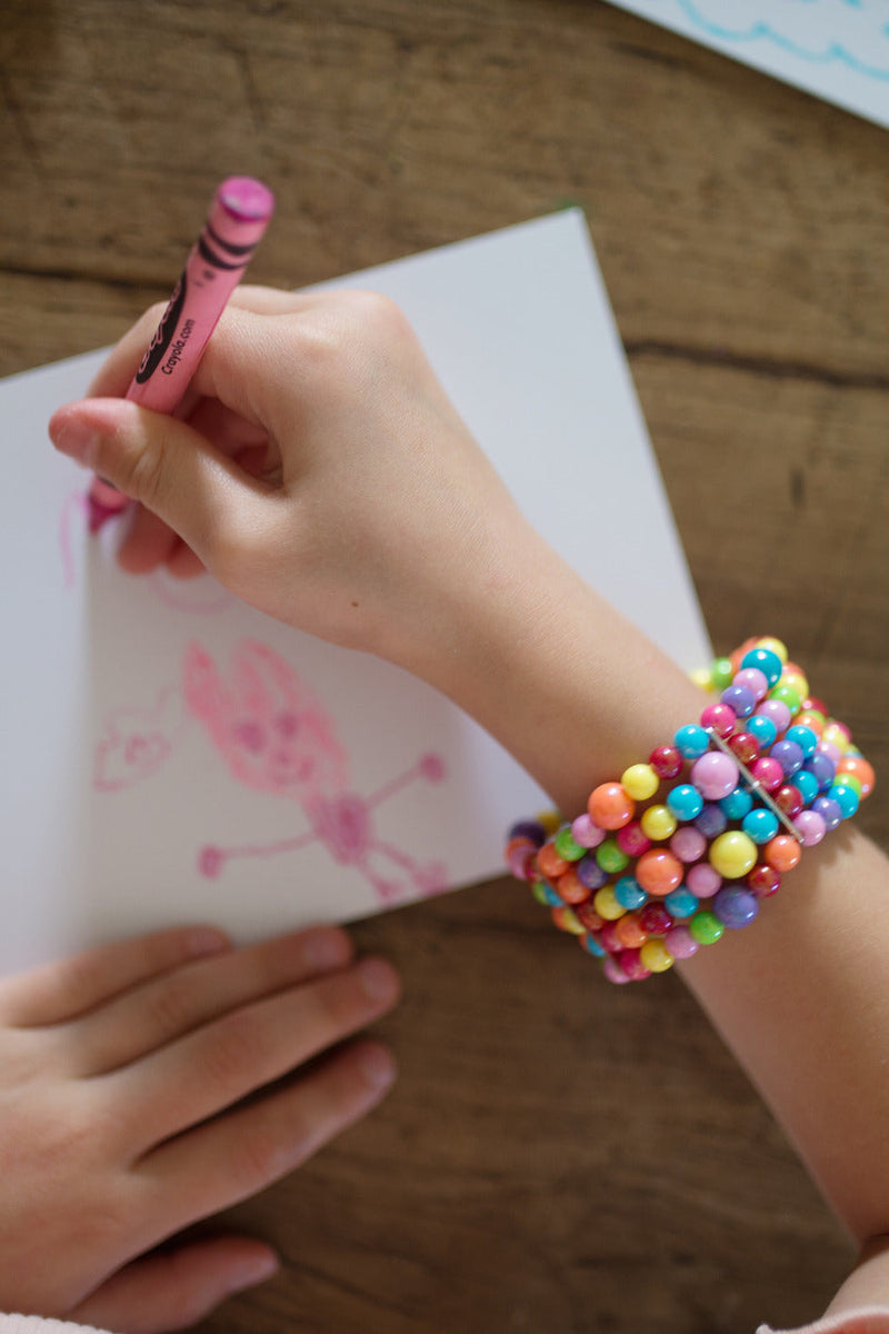 Child wearing colorful beaded bracelet while drawing with pink crayon on paper