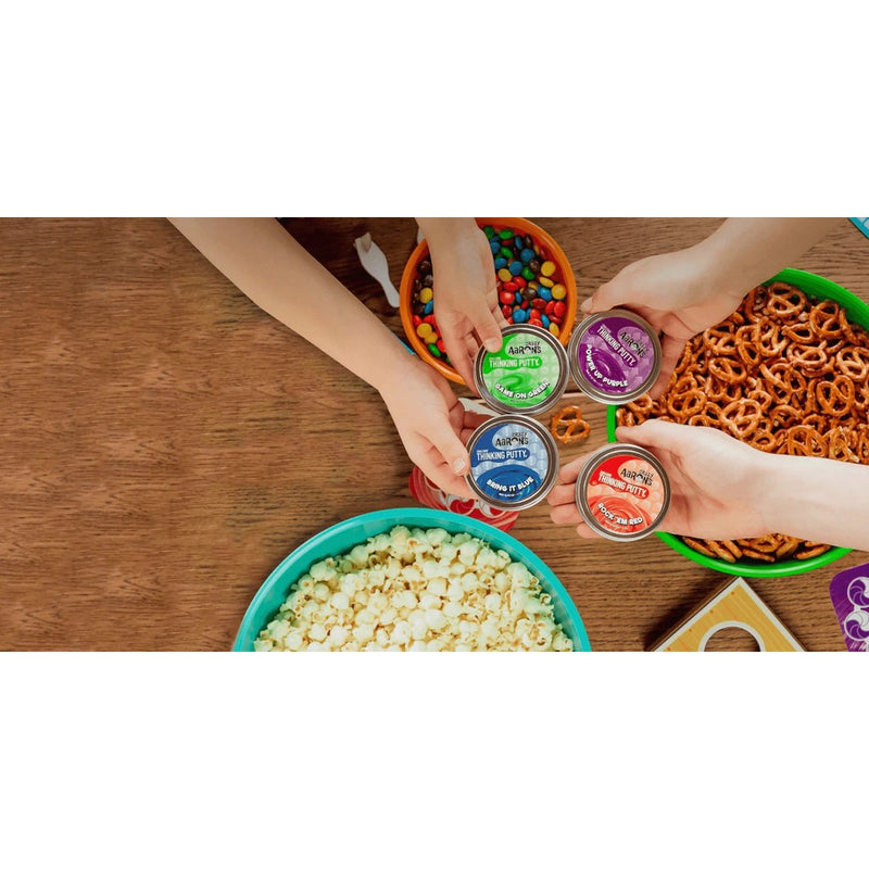 Four mini tins of Crazy Aaron's Thinking Putty displayed with hands and snacks on wooden table