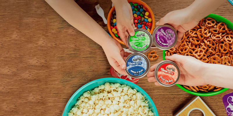 Four mini tins of Crazy Aaron's Thinking Putty displayed with hands and snacks on wooden table
