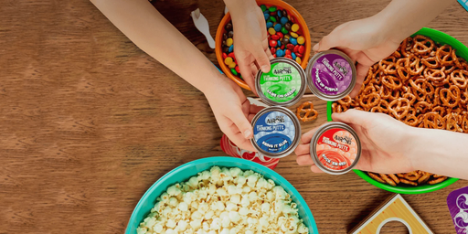 Four mini tins of Crazy Aaron's Thinking Putty displayed with hands and snacks on wooden table