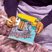 Person holding FlowArt Color-to-Reveal spiral notebook with vibrant illustrated cover on purple bedding