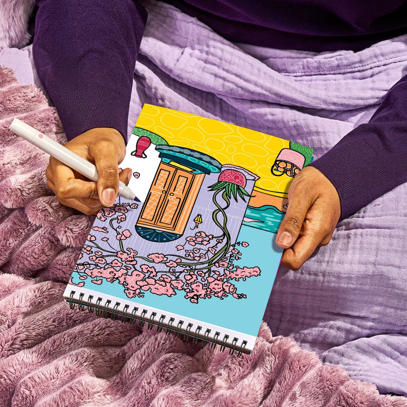 Person holding FlowArt Color-to-Reveal spiral notebook with vibrant illustrated cover on purple bedding