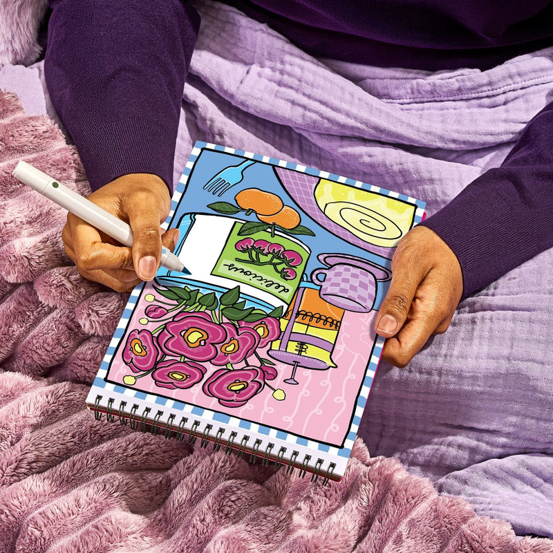Person sitting on purple bedding holding FlowArt Fragrant Blooms color-to-reveal spiral notebook with marker