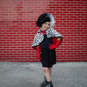 Child wearing Cruella De Vil costume with spotted cape, black wig, and red gloves
