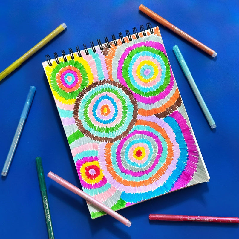 OOLY Rainbow Sparkle glitter markers displayed around a colorful spiral circle artwork in a sketchbook
