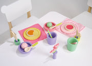 Colorful wooden toy lunch set from Mentari with plates, cups, food items on white table with chairs