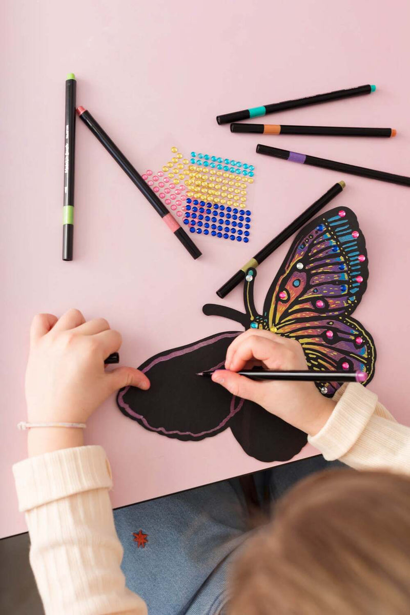 Child using metallic markers to color butterfly scratch art with colorful gem stickers